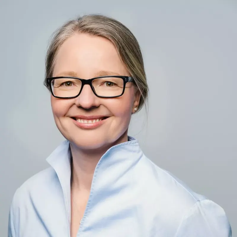Porträt einer lächelnden Frau mit Brille in hellblauer Bluse vor hellem Hintergrund